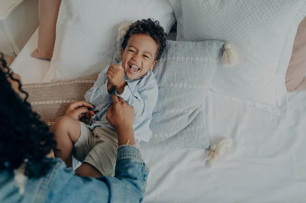 Sweet little boy kid laughing loudly and smiling while his loving mom tickling him on bed with lot of soft pillows, mother telling really funny story to son while spending happy time together at home