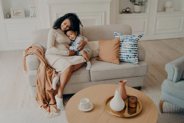 Loving african pregnant mom tenderly embracing little son while sitting on cozy couch next to coffee table in living room, spending leisure time together at home during weekend. Motherhood concept