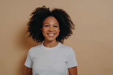 Güzel, genç Afrikalı bir kadın kameraya neşeyle gülümsüyor ve mutluluğunu gösteriyor. Stüdyoda renk duvarının karşısında duruyor. Siyah tenli, kıvırcık saçlı, beyaz dişlerini gösteren bir kadın.
