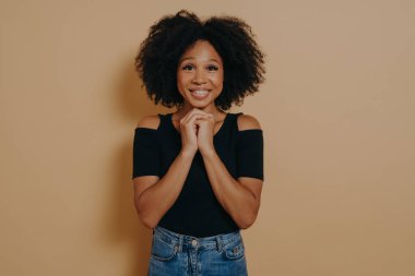 Karışık ırktan genç bir kadın dua jestinde el ele tutuşuyor, gözleri açık gülümsüyor koyu bej arka planda izole bir şekilde fotokopi çekiyor. Stüdyoda poz veren Afrikalı kadın, umut ve olumlu duygu kavramı