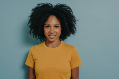 Stüdyoda mutlu bir gülümsemeyle poz veren neşeli, genç, melez, kıvırcık saçlı bir kadın portresi. Koyu tenli kadın sarı tişört giyiyor, neşeyle gülümsüyor, beyaz düz dişlerini gösteriyor.