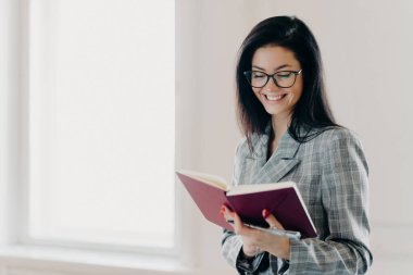 Tatmin olmuş esmer kadın mutlu bir ifadeye sahip. Çalışma planını not defterine yazıyor. Kırmızı manikür resmi kıyafet kayıtları.