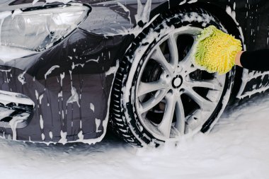 Cropped shot of black automobile washed by service worker at carwash. Vehicle covered with foam bubbles. Focus on wheel.