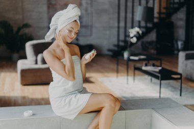 Gülümseyen genç bir kadın sağlıklı, parıldayan tenine dokunur, evde günlük güzellik prosedürleri yapar, bir şişe losyon tutar, yüz kremi sürer, banyo havlusuna sarılır, sıcak ev içi pozlar verir.