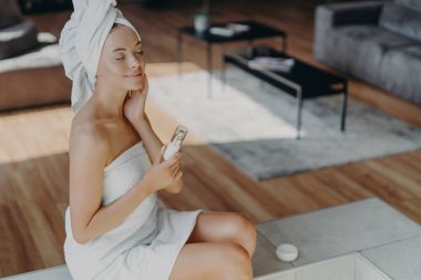 Tatmin olmuş, rahat, güzel bir kadın, gözlerini kapatır ve teninin yumuşaklığından zevk alır, duştan sonra güzellik tedavileri yaptırır, bir şişe losyon tutar, evde poz verir, rahatlık ve haz hisseder.