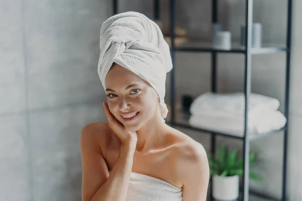 Beautiful European woman with makeup touches skin, has minimal makeup, has healthy glowing skin, wrapped in bath towel, enjoys rest at home. Spa woman poses in bathroom. Beauty, wellness concept