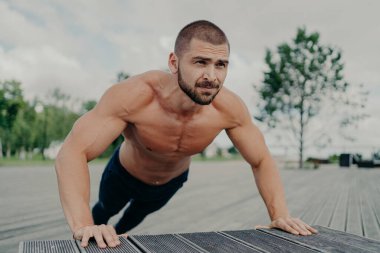 Aktif kaslı adam egzersiz yapıyor üstsüz dışarı çıkıyor, parkta egzersiz yapıyor, kalın sakalı var, alt pozisyonda duruyor. Motive olmuş vücut geliştirici düzenli olarak egzersiz yapar. Sporcu üst gövdeyi eğitir