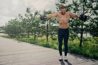 Atletik güçlü fitness adamı halterlerle çalışır, güç ve enerji gösterisi yapar, aktif bir hayat sürer, güç gösterir, motivasyon gösterir, pazı kaldırır, dışarıda fiziksel egzersiz yapar.