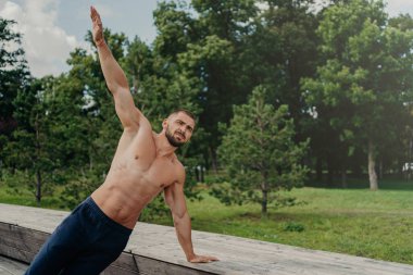 Güçlü genç adam tek kolunda yan tahtada duruyor, dengeyi buluyor, ağaçların yanındaki parkta poz veriyor, dışarıda yoga yapıyor, aktif sağlıklı bir yaşam tarzı sürüyor, kaslı güçlü bir vücudu var. Motivasyonlu vücut geliştirici