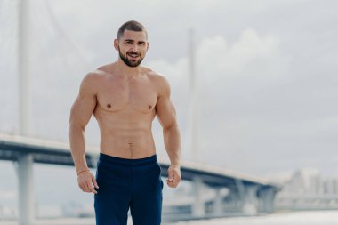 Çıplak gövdeli neşeli sakallı adam kameraya sevinçle bakar, pantolon giyer, nehir köprüsüne karşı poz verir, sağlıklı bir hayat sürer, düzenli antrenmanlardan sonra kaslı bir vücudu vardır.