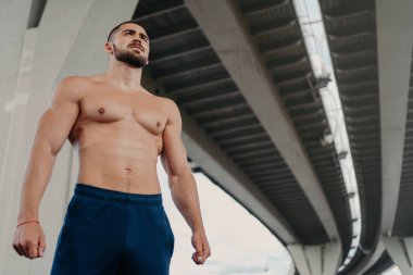 Kendine güvenen sakallı yakışıklı Avrupalı bir adamın güçlü bir vücudu var. Uzağa bakıyor, aktif bir yaşam sürüyor, köprü altında poz veriyor. Genç üstsüz vücut geliştirici..