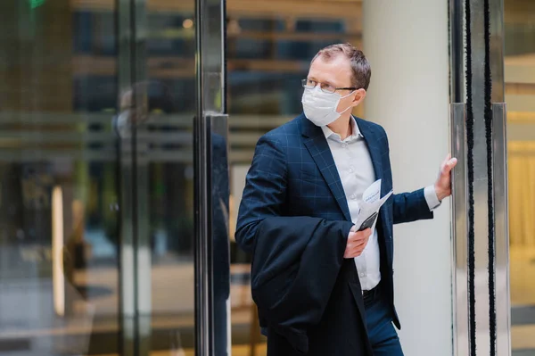 Respiratory protection, coronavirus, dangerous disease. Serious man stands in doors of office building, wears medical mask, holds modern cellphone, newspaper to read article about covid-19 treatment