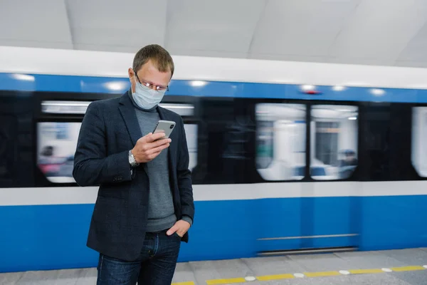 Male passenger wears face mask poses at platform, waits for train, commutes by underground, concentrated in smartphone device, reads news online. Virus awereness in public place. Coronavirus outbreak