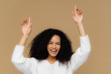 Güzel koyu tenli kadın modeli yatay çekim dişli gülümseme vardır, eller yükseltir, puan kadar, overjoyed hissediyor, kıvırcık saçlı, beyaz kazak giyer, kahverengi arka plan üzerinde izole. Kadın eğleniyor