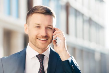 Müreffeh başarılı tıraşsız işadamı açık atış resmi giysiler giyiyor, cep telefonu üzerinden iş ortağı ile hoş konuşma vardır, gelecekteki toplantı, pozlar arka plan bulanık karşı tartışmak