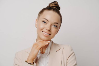 İş konferansında bej takım elbiseli ve çörekli güzel bir kadın iyi haberler dinliyor, elleri gri arka planda izole edilmiş bir şekilde kameraya bakarken mutlu ve rahat oluyor.