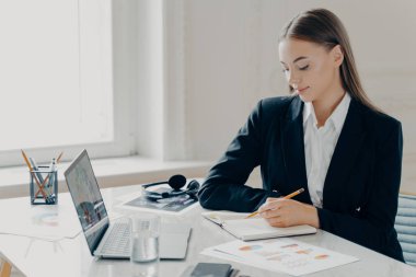 Siyah takım elbiseli, gülümseyen, beyaz, minimalist arka planda penceresi olan beyaz bir masanın yanında oturan beyaz bir kadının yan portresi..