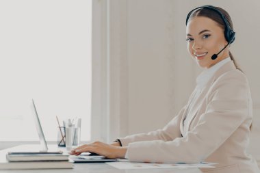 Genç bayan çağrı merkezi ajanının mikrofon danışmanı müşterisiyle kulaklık takması ve bilgisayar kullanması göz önüne alındığında, iş kadını resmi takım elbise giyerek çevrimiçi toplantıda yer alıyor.