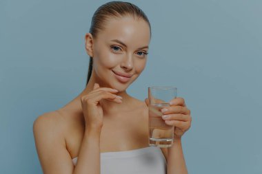 Healthy and hydrated skin. Young charming woman holding glass of pure clean water and touching moisturized facial skin, smiling at camera, posing on blue background. Skincare and healthcare concept