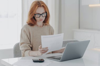 Gözlüklü, gözlüklü genç bir kadın evde bütçesini hesaplıyor, dizüstü bilgisayarla masaya oturuyor, kağıt vergileri tutuyor ve mali bilgileri okuyor, aile bütçesini yönetiyor.