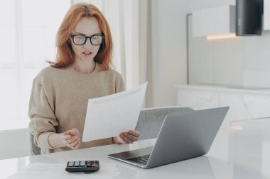 Zencefilli profesyonel kadın ekonomistin kapalı çekimleri, kağıt tutulan evlerdeki finansmanı hesaplıyor. Finansal rapor, modern dizüstü bilgisayar ve hesap makinesiyle çevrili beyaz masaüstünde duruyor.