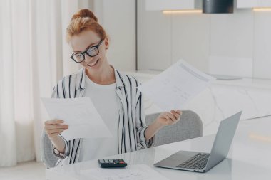 Sıradan giyinmiş, heyecanlı kızıl saçlı bir kadın evinde oturuyor, ev faturalarını kontrol ettikten sonra mutlu oluyor, elinde banka tebligatı ile belgeleri tutuyor ve gülümsüyor, dizüstü bilgisayar ve hesap makinesi kullanıyor.