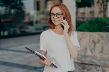 Mutlu kızıl kadının yatay görüntüsü gözlük takıyor. Günlük beyaz tişört giyiyor. Telefon görüşmeleri yapıyor.