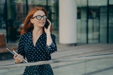 Düşünceli kızıl saçlı kadın gözlük takıyor, benekli elbise giyiyor telefon görüşmeleri yapıyor, ofis arka planındaki bulanık yerlere bakıyor çalışmak için gerekli şeyleri taşıyor. Yaşam biçimi