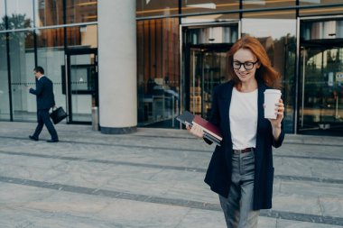 Güzel kızıl saçlı iş kadınının açık havada çekilmiş fotoğrafı, not yazmak için taşınabilir dijital tablet defter taşıyor ve ofis binasının yakınındaki iş toplantısından dönerken harcanabilir kahve yürüyüşleri yapıyor.