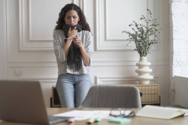 Ateşini ölçen, evden uzakta çalışırken yüksek ateş ölçen atkısı olan, serbest çalışan genç hasta kadın termometreye endişeli yüz ifadesiyle bakıyor, gripten muzdarip mutsuz kadın.