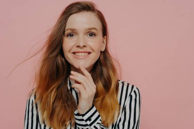 Hoş, gülümseyen, kızıl saçlı, resmi çizgili, siyah beyaz bluzu olan, açık pembe stüdyo arka planında duran, çenesine dokunurken gülümseyen genç bir kadının fotoğrafı. Mutluluk kavramı