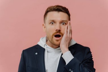 Şok olmuş, sakallı, ceketli, kapüşonlu, kırmızı saçlı, çenesi düşüp pembe duvarın üzerinde tek başına dikilirken kameraya bakan yakışıklı bir adam. İnsan duyguları kavramı