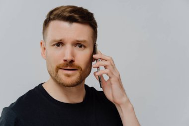 Cep telefonuyla konuşan kafası karışmış bir gencin portresini yakın çekin ve kötü haber aldıktan sonra mutsuz hissedin, endişeli bir yüz ifadesiyle kameraya bakın, gri stüdyo duvarına karşı durun.