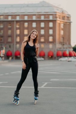 Full length shot of active smiling woman enjoys extreme sport rides on rollerblades dressed in black comfortable clothes stands outdoors learns rollerskating. People leisure recreation concept