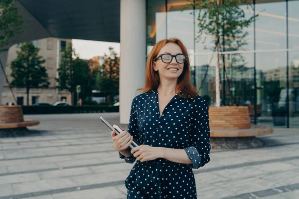 Successful woman entrepreneur carrying laptop notebook while standing on city street, looking aside with smile, smiling female freelancer with digital device in hand enjoying beautiful day outside