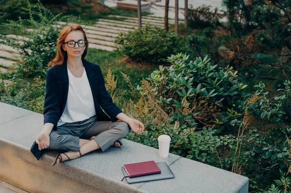 Peaceful calm redhead business woman doing yoga with closed eyes outdoor, relaxing in lotus pose during remote work in park, female relieving stress while working on laptop outside. Meditation concept