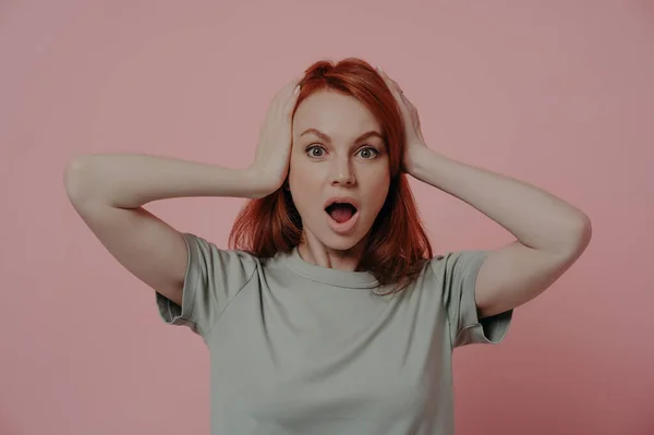 Indoor shot of shocked scared woman keeping head in hands, staring at camera with bugged eyes from surprise or wonderment and dropped jaw, being in shock while posing isolated over pink background