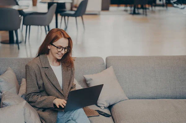 Beautiful happy female employee in casual clothes sitting on comfortable couch in office break room, using laptop to browse web, smiling and feeling satisfied, furniture in blurred background