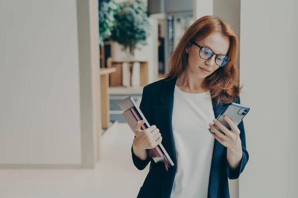 Redhead business woman with serious look in eyewear using cell phone, checking email or newsfeed of social network account, holding laptop and notebook, looking at smartphone while standing in office
