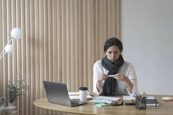 Unhappy sick young hispanic woman office worker in scarf around neck looking at electronic thermometer while sitting at her workplace, ill female suffering from covid19 or flu symptoms at work