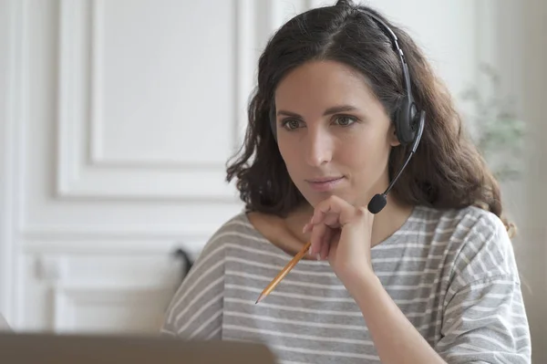 Focused young Italian entrepreneur lady wearing headset attentively listening educational webinar looking at laptop screen, getting remote online office trainings at home. Telemarketing concept