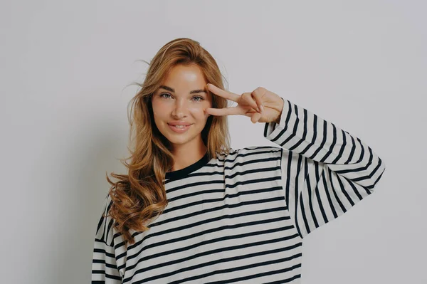 Body language concept. Portrait of young joyful optimistic girl in striped blouse long sleeve showing V-sign or peace gesture with fingers near eye and looks at camera positively smiling