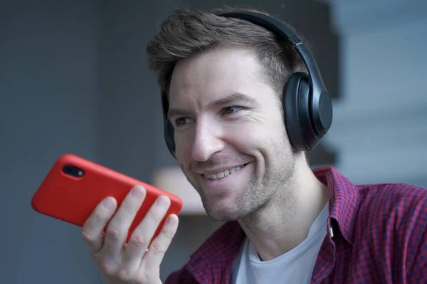 Close up of cheerful young german man in wireless headphones holding smartphone in hand with microphone close to his face during mobile conversation with friend, recording audio message