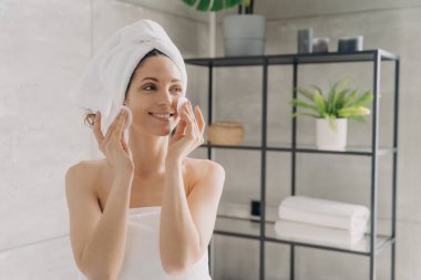 Güzel İspanyol kız yüz derisini pamuk pedlerle temizliyor. Havluya sarılı gülümseyen kadın banyodaki duştan sonra makyajını siliyor. Cilt günlük rutin, doğal kadın güzellik konsepti.