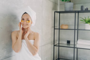 Göz altı nemlendirici hidrojel bantlarıyla gülümseyen güzel bir kadın. Cilt tedavisi sırasında beyaz banyo havlusu takan güzel bir kadın..