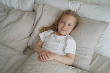 Yatak odasındaki rahat yatakta yorganla kaplı küçük sakin bir kız çocuğu. Tatlı beyaz çocuk uyandıktan sonra rahat bir yatakta yumuşak yastığın üzerinde dinleniyor. Sağlıklı çocuk uyku konsepti.