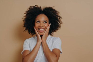 Neşeli koyu tenli genç bir kadın mutlu, kameraya gülümsüyor ve iki eliyle yanaklarına dokunuyor, kaygısız beyaz tişörtlü, pozitifliğini ifade eden, bej arka planda izole edilmiş poz veren Afrikalı bir kadın.
