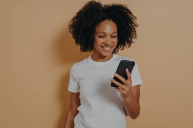Mutlu gülümseyen Afrikalı kız öğrenci cep telefonu kullanıyor ve gülümsüyor, internette olumlu haberler okuyor veya internette erkek arkadaşıyla sohbet ediyor, kum rengi duvara karşı poz verirken modern akıllı telefonu tutuyor.