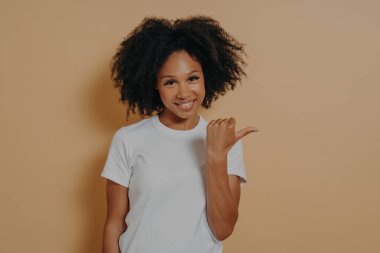 Genç, koyu tenli, kıvırcık saçlı, baş parmağıyla işaret eden kadın. Terfi için boş bir alan ya da bej duvarın üzerinde izole edilmiş, pozitif yüz ifadesi olan bir reklam gösteriyor.
