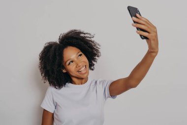 Neşeli, kıvırcık saçlı, modern cep telefonunda selfie çeken, beyaz stüdyo arka planında izole edilmiş beyaz tişörtlü, mutlu bir şekilde gülümseyen genç Afrikalı kadın.